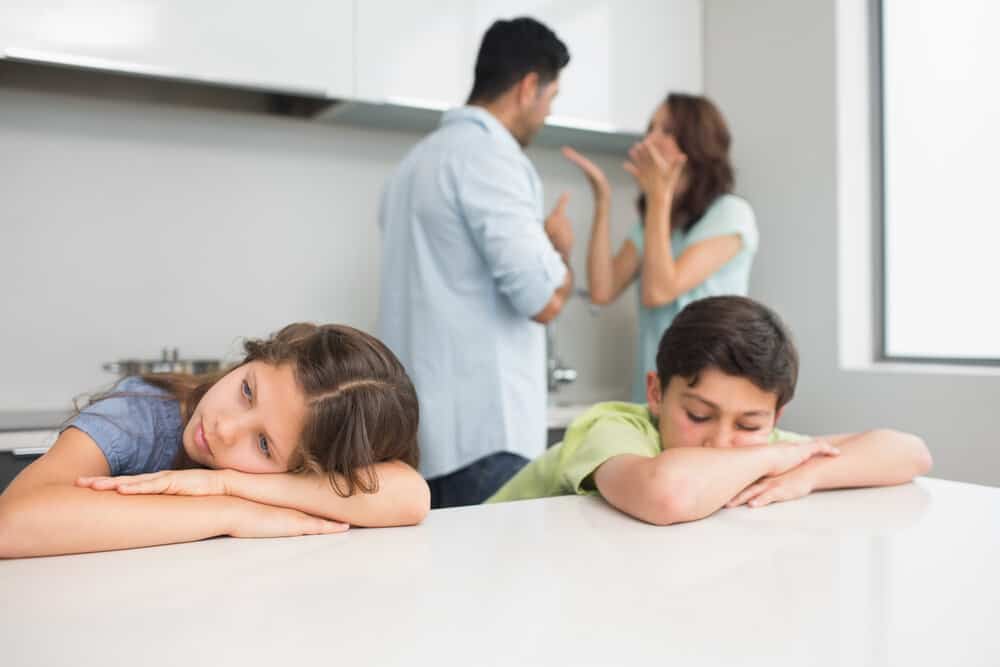 Two children lay with their heads on a table while parents fight behind them.